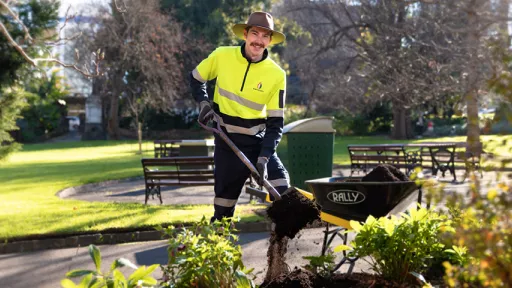 Gardener wearing hi-vis top and hat shovels compost from a wheelbarrow to a garden bed in a park.