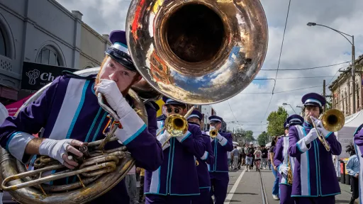 Members of brass band wearing purple and white uniforms walk along Glenferrie Road. 