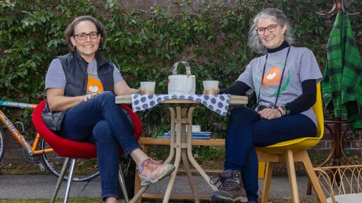 Two volunteers sit in garden beside table. In background are a bike, a coat rack, jacket and basket.