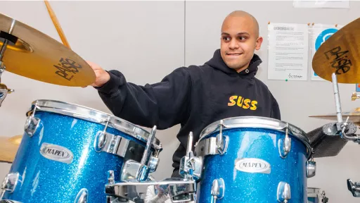 Smiling student wearing blue hooded jumper sits behind drum set playing blue drums and brass cymbals.