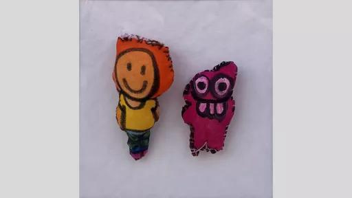 Two small dolls. The left doll looks like a person smiling with orange hair, yellow t-shirt, and khaki pants. The right doll is a pink monster with 3 big buck teeth.