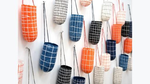 Image of over 20 paper vessel sculptures, depicting danala or dillybags, pined to a wall. Each dilly bag sculpture has a grid pattern painted on the surface. The colour variations of the dillybag sculptures are white lines on blue background, white lines on orange background, white lines on black background, black lines on white background, and orange lines on white background. 