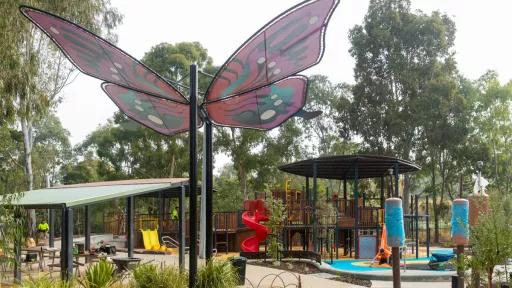 A playground with a prominent butterfly sculpture, constructed from fabric in shades of pink and green, with a metal framework. The background features a substantial wooden playground structure, complete with a slide and climbing apparatus. Adjacent to this is a picnic shelter, offering a table and seating. The playground is surroundedby trees.