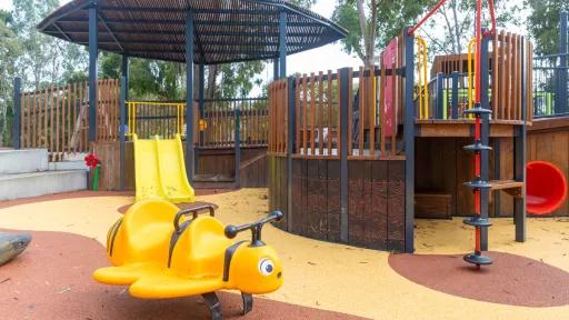 A yellow bee-shaped playground ride is featured on a brown and yellow rubber safety surface. Adjacent is a wooden playground structure, complete with a red and yellow slide, a climbing wall, and a red tunnel. The entire play area is sheltered by a black wooden roof with a brown and yellow wood-patterned side.