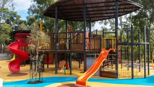 A multi-level play structure with a wooden base and a black metal roof. The structure includes two slides, one red and one orange, as well as a variety of climbing components and climbing walls. The play area is surrounded by trees and shrubs, and the ground is covered with rubber mulch in a variety of colors.