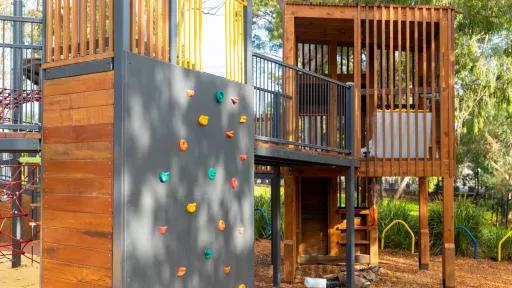 A wooden climbing frame with a grey climbing wall and a platform made of wood with a wooden railing. There are a variety of colored hand and foot holds on the climbing wall.