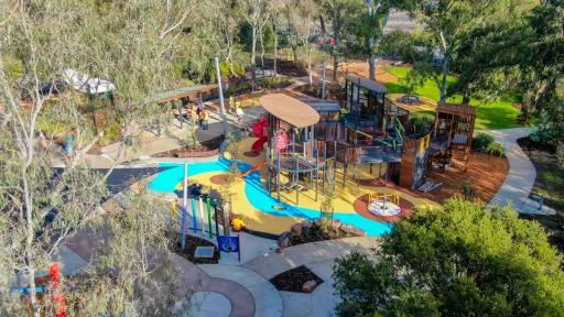 The image depicts an aerial view of a playground. Various play structures are visible, such as a large wooden unit with slides, a climbing wall, and a swing set. Adjacent is a blue and yellow play area with a meandering path. The playground is nestled amidst trees and grass, with individuals walking along the paths and engaging with the equipment.