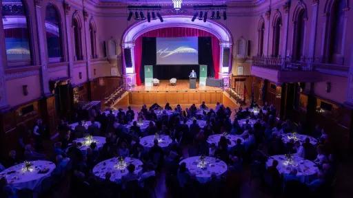 Aerial perspective of a large hall. A stage is at the far end, with a speaker addressing the assembly from the lectern. The hall is filled with round tables occupied by individuals. The hall has a purple glow.