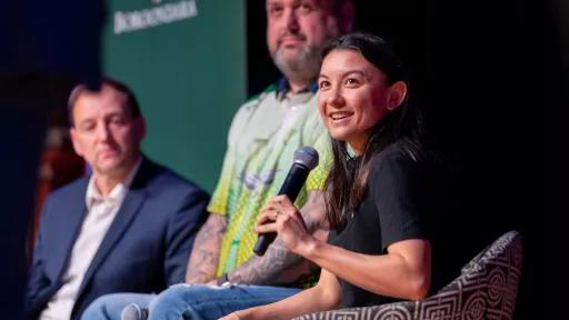 A woman is seated on a stage, microphone in hand, smiling at the camera. Two men sit beside her in the background.