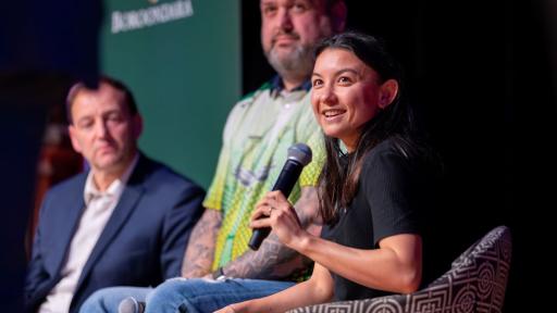 A woman is seated on a stage, microphone in hand, smiling at the camera. Two men sit beside her in the background.