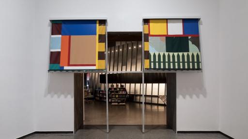 Photograph of two artworks in a gallery. The artwork consists of semi-abstract shapes printed onto fabric and turned into window awnings, built for external windows. They are both positioned above an open doorway so that people can walk underneath them. The shapes in the patterned fabric resemble windows and fences and use various shades of blues, oranges, yellows, greens, browns, and white. 