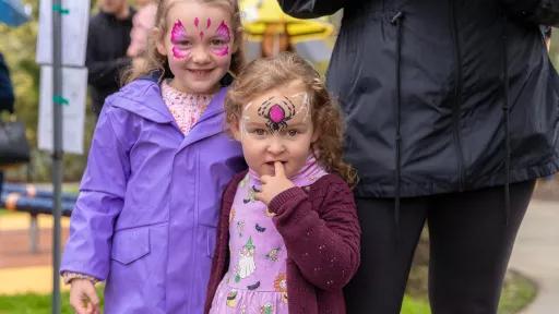 2 small children standing with an adult. 1 child is wearing a purple raincoat and has butterfly face paint. the other is wearing a purple dress and cardigan and gas spider facepaint.