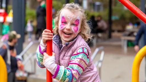 child in a woolen jumper and pink vest plays on the playground. she is smiling and has butterfly facepaint.