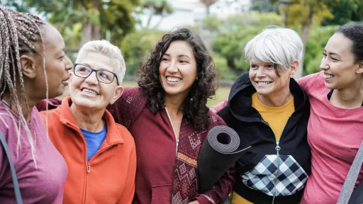 Five women are standing outside and smiling. The women are all wearing casual clothes and are standing with their arms around each other. They are all different ages and ethnicities.