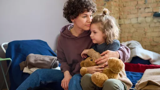 A woman sits on a bed, hugging a young girl who is holding a teddy bear. The woman’s hand rests on the girl's back in a comforting manner. There are blankets on the bed in the background.