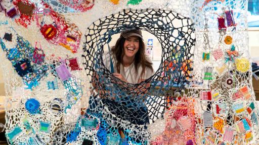 A woman is smiling directly at the camera looking through a large brightly coloured crocheted blanket 