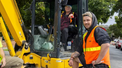 Two road workers in hi-vis tops stand next to yellow digger with third worker in digger cab.