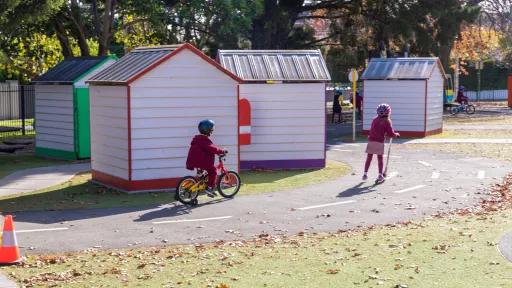 two children one on a bike, the other on a scooter riding along the road around buildings