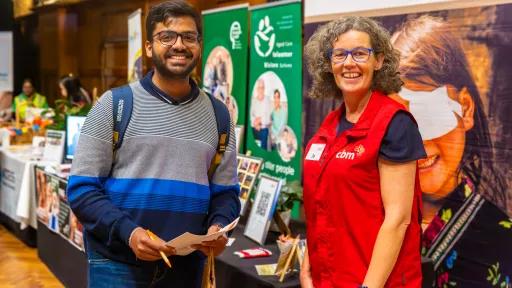 2 people standing at a volunteer expo booth. one is holding a pencil and paper.