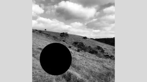 The black-and-white photograph shows rolling grassy hills with scattered trees under a textured, cloud-filled sky. The terrain slopes upward, with vegetation varying in density. In the foreground, a large, solid black circle contrasts with the rough grass. Scattered trees are dispersed across the hills, with one near the crest standing against the sky. The background features more hills and a fading tree line.