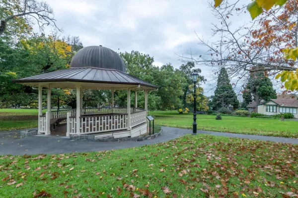 Rotunda with cream-white balustrades and grey dome-shaped roof, surrounded by a footpath and grass area. There is a lamp post in the centre and fallen leaves on the ground.