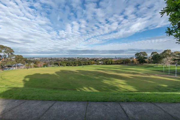 Oval-shaped grass field surrounded by two large grass and brick steps. There are trees and houses are in the distance and a footpath in the foreground.