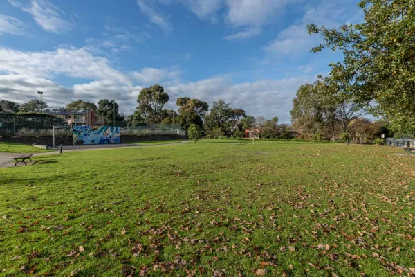 Large grass area with fallen leaves, with trees to the right and in the distance. There is a painted mural, a park bench and a footpath to the left. 