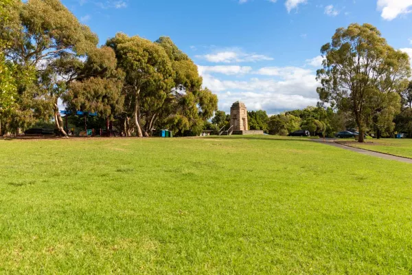 Large grass area with several large trees in the distance. There is a footpath to the right leading to a concrete memorial structure.