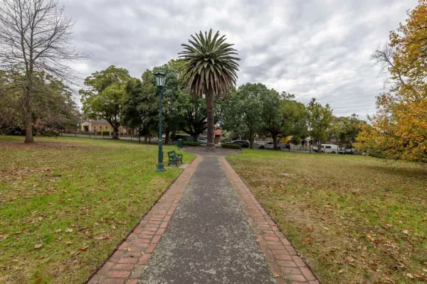 Straight walking path with grass and fallen leaves on either side, leading to a small circular feature with a tall tree. There is a lamp post and park bench to the left of the path.