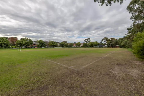 Corner view of soccer pitch under grey sky with goals at each end and medium trees on the horizon. There is a large bare dirt patch in the foreground.