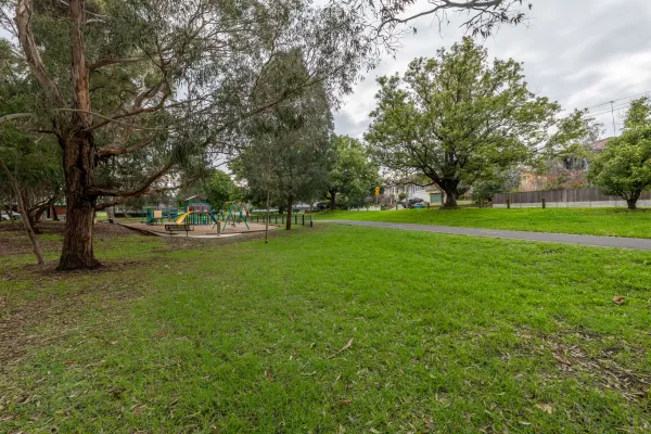 Small grass area with distant playground shaded by several tall trees. There is a walking path and fence to the right.