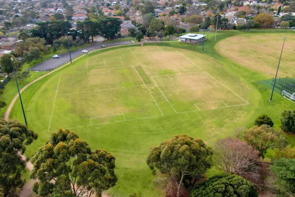 Aerial view of oval sportsground with various square line markings on its surface and light posts around its edge. There are cricket nets to the bottom right.