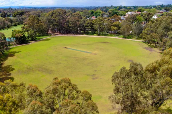 Aerial view of oval-shaped grass field with cricket pitch. There is a thick layer of trees around the oval and houses to the far right.