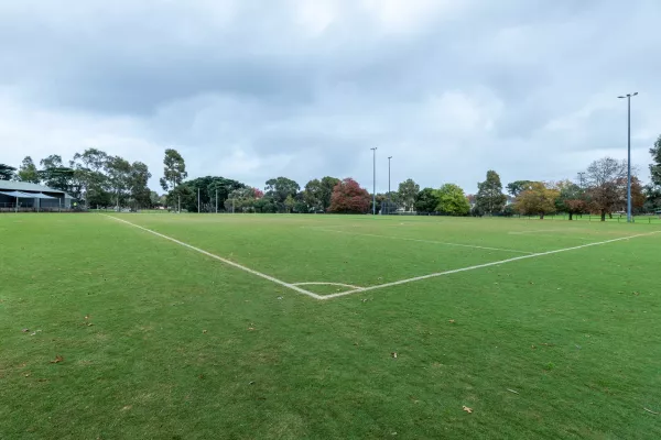 Corner view of soccer pitch with no goals at near end and AFL goal posts at far end. There is a one-storey building at the distant left and five visible light posts.