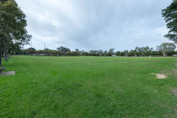 Large grass field with dirt walking track to the right and a long row of medium trees and park benches to the left. There are soccer goals in the distance.