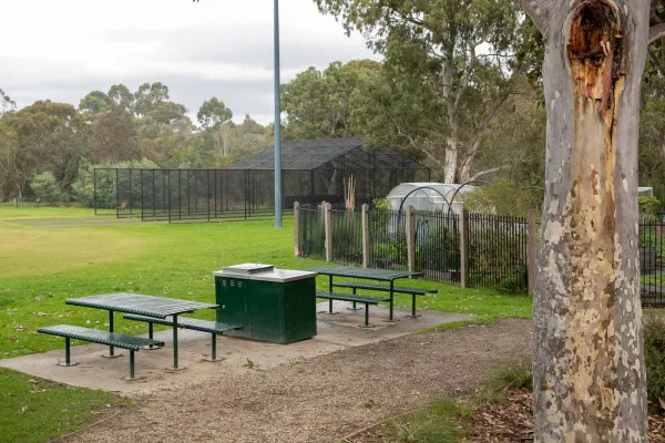 Barbecue and two sets of tables and chairs between a grass field and a dirt path. There is a tree trunk to the near right, a fenced-off nursery in the background and cricket nets in the distance.