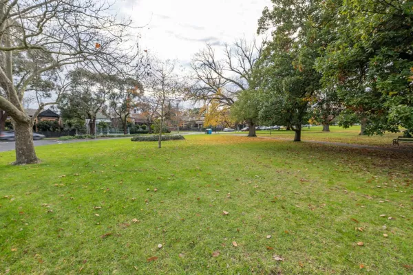 Large grass area with fallen leaves and scattered mature trees. There is a park bench and walking path to the right and a line of houses in the distance.