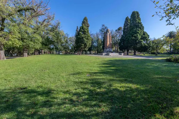 Area of lush grass in sunshine with concrete memorial structure and tall trees at the far end. There are shadows in the foreground.