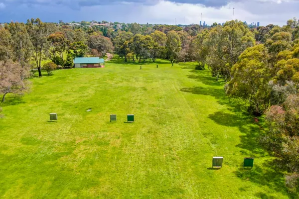 Large rectangular grass area surrounded by a thick layer of trees. A small single-storey building is at the far end. There are five archery targets in the foreground.