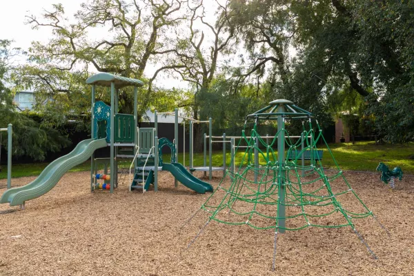 Playground with swings, rope feature and a small climbing feature with two slides. There are large trees in the background.