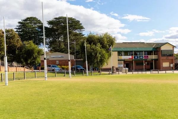 Part of an oval grass field with tall trees and white goalposts to the right. There are two house-sized buildings on the field edge.