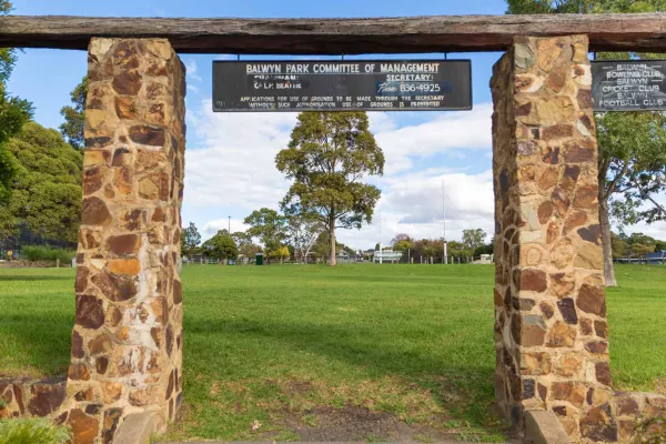 Rectangular entrance structure for grass field, with two brick columns, two information signs and a wooden plank. There are tall trees are in the distance.