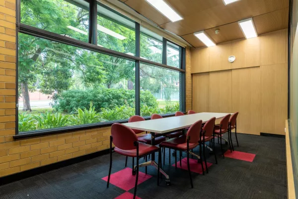 A small room with a long rectangular table surrounded by red chairs. A large window overlooks the trees outside 