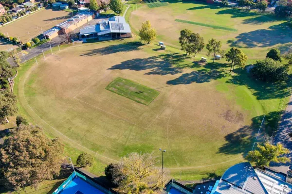 An aerial view of a green sports oval surrounded by trees. A building sits on the edge of the oval. a second sports oval sits beside it. 